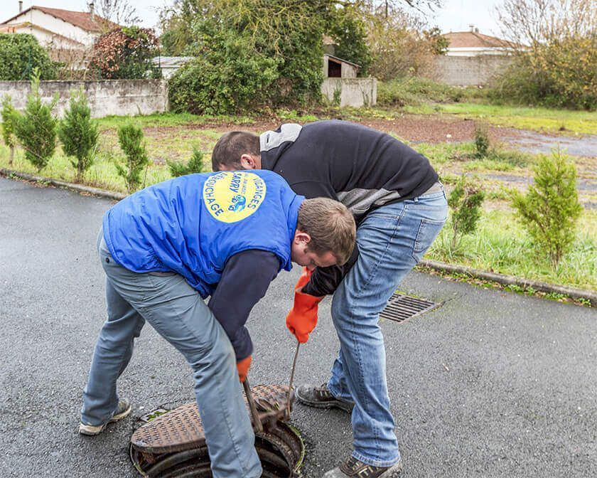 Deux personnes ouvrent une plaque d'égout sur une rue asphaltée. L'une d'elles porte un gilet bleu.