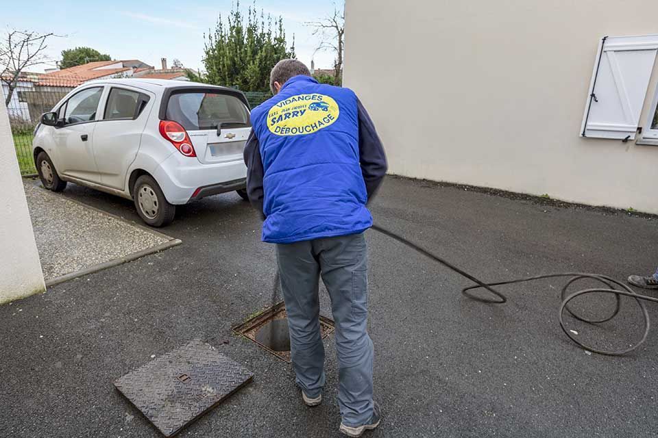 Un homme en gilet bleu travaille sur un drain dans une allée près d'une voiture blanche et d'une maison.