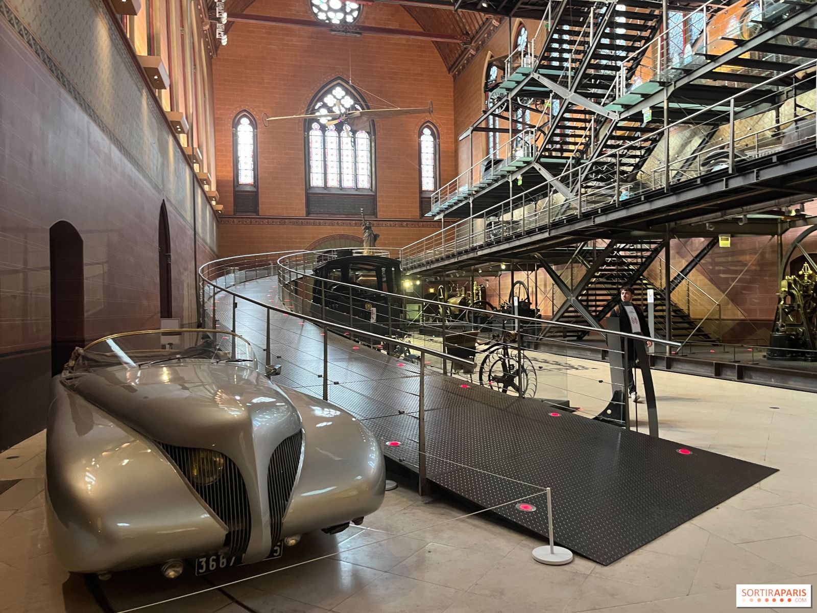 Silver streamlined car on display in a large hall with metal staircase and gothic window.