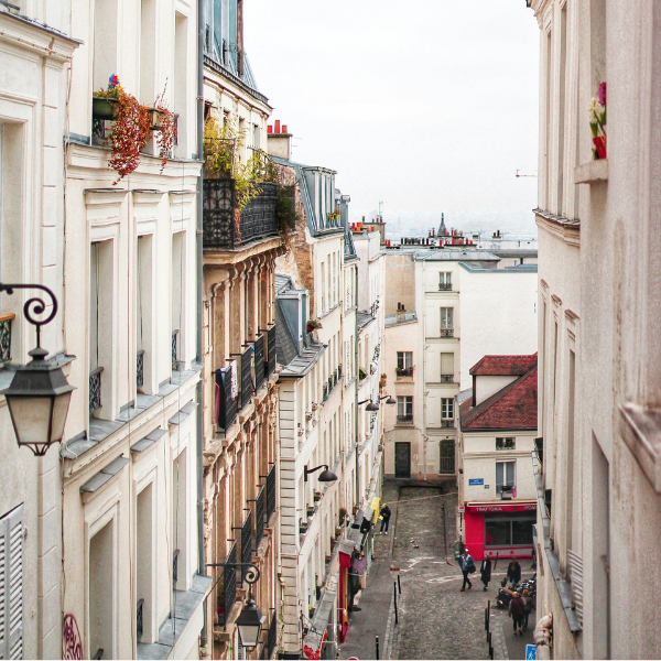 Narrow street lined with beige buildings, ornate balconies, and a red-roofed building. People walk.