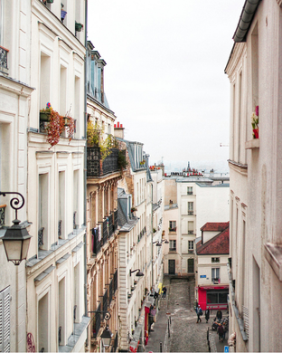 Narrow Parisian street lined with tall, cream-colored buildings. Street slopes downward; pedestrians and shops visible. 