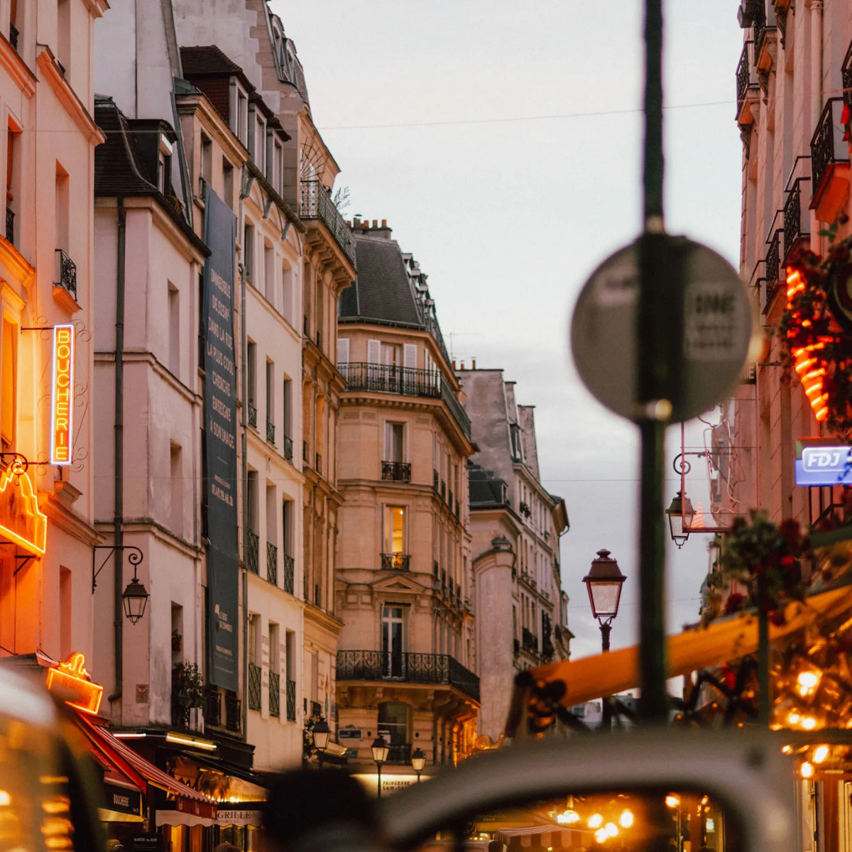Narrow Parisian street lined with tall, cream-colored buildings. Street slopes downward; pedestrians and shops visible. 