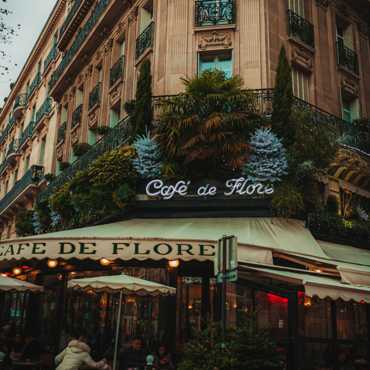 Narrow Parisian street lined with tall, cream-colored buildings. Street slopes downward; pedestrians and shops visible. 