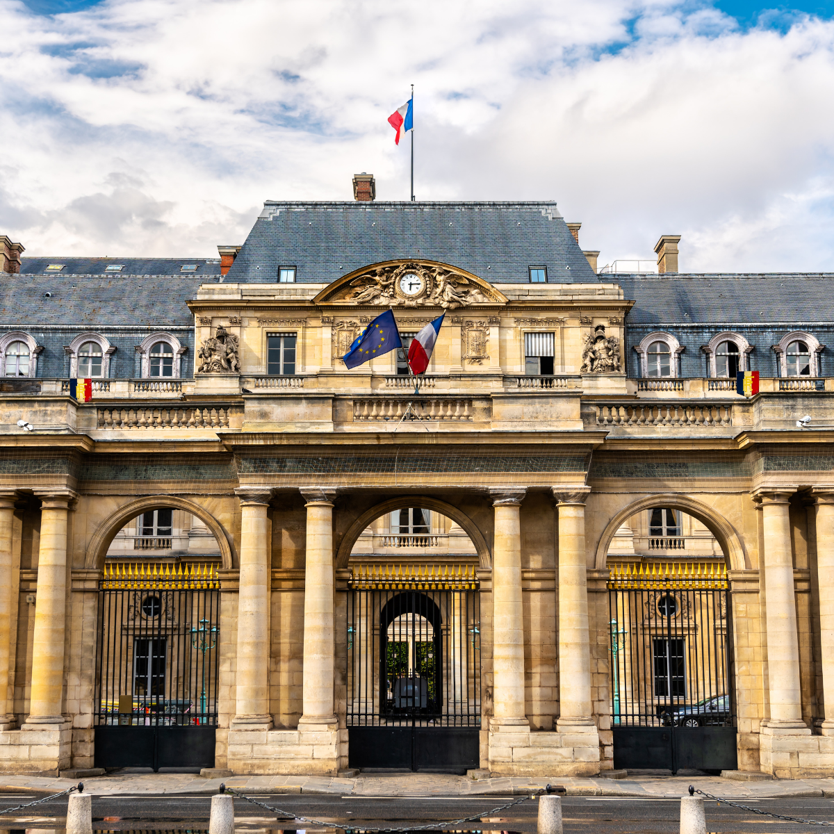 Narrow Parisian street lined with tall, cream-colored buildings. Street slopes downward; pedestrians and shops visible. 