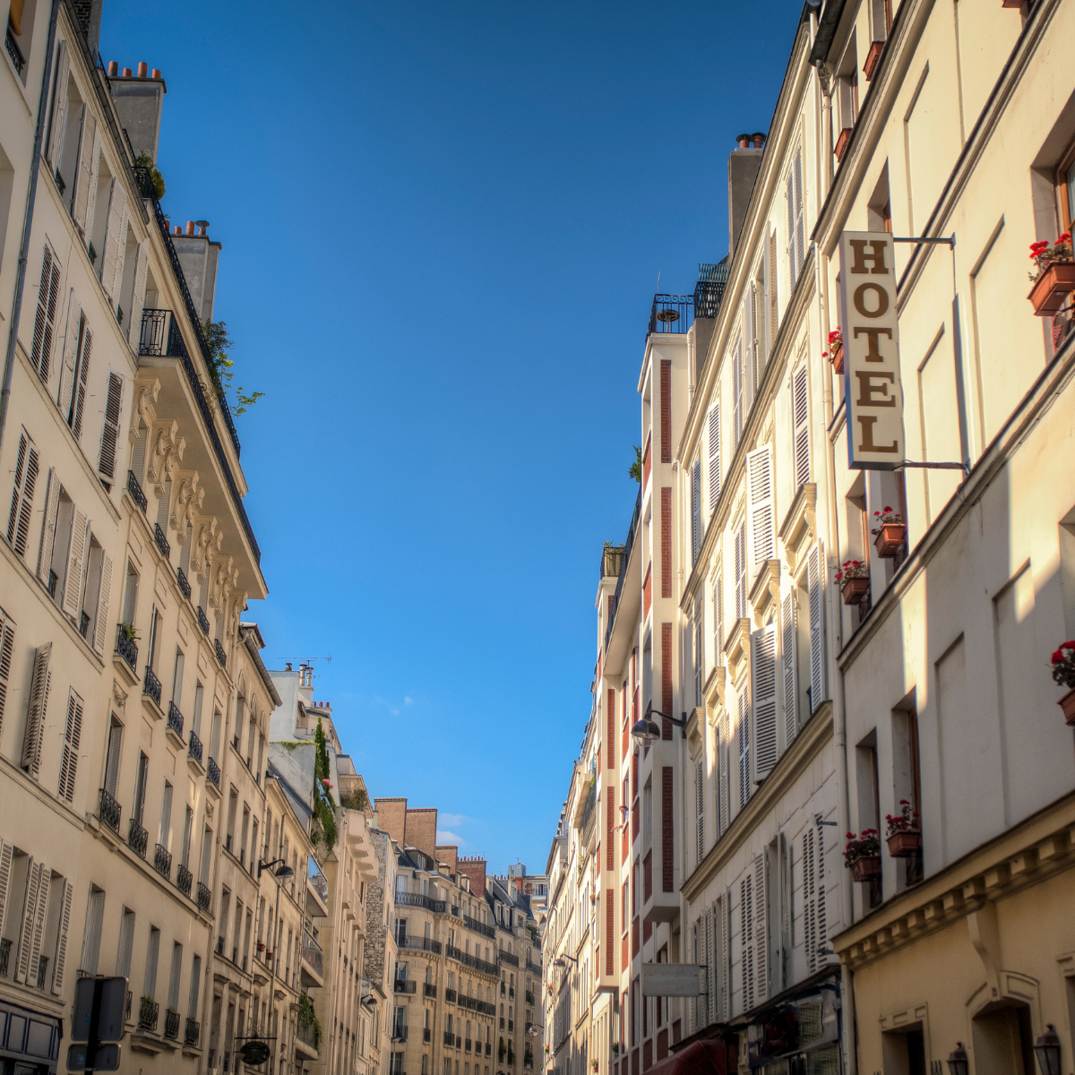 Narrow Parisian street lined with tall, cream-colored buildings. Street slopes downward; pedestrians and shops visible. 