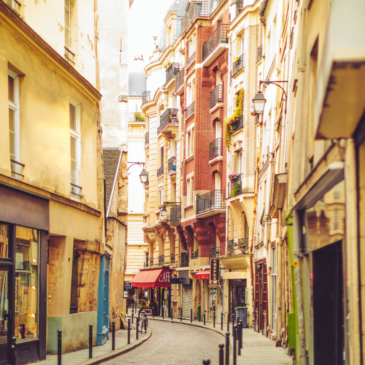 Narrow Parisian street lined with tall, cream-colored buildings. Street slopes downward; pedestrians and shops visible. 