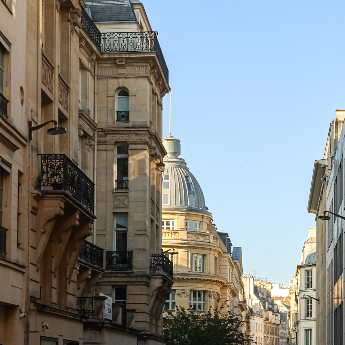 Narrow Parisian street lined with tall, cream-colored buildings. Street slopes downward; pedestrians and shops visible. 