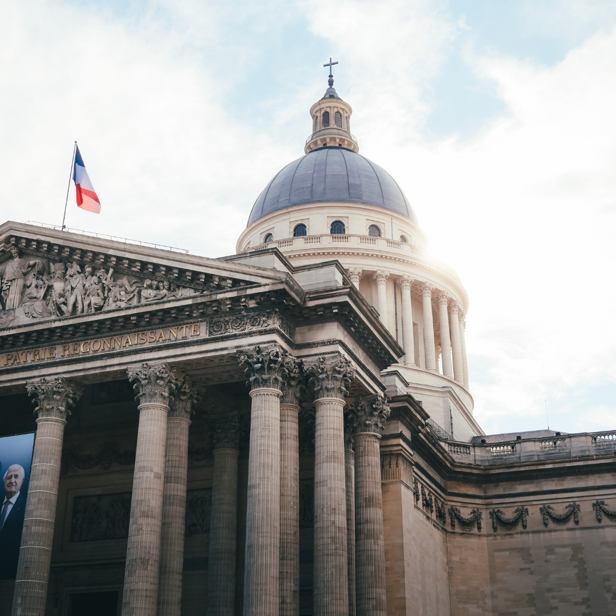 Narrow Parisian street lined with tall, cream-colored buildings. Street slopes downward; pedestrians and shops visible. 