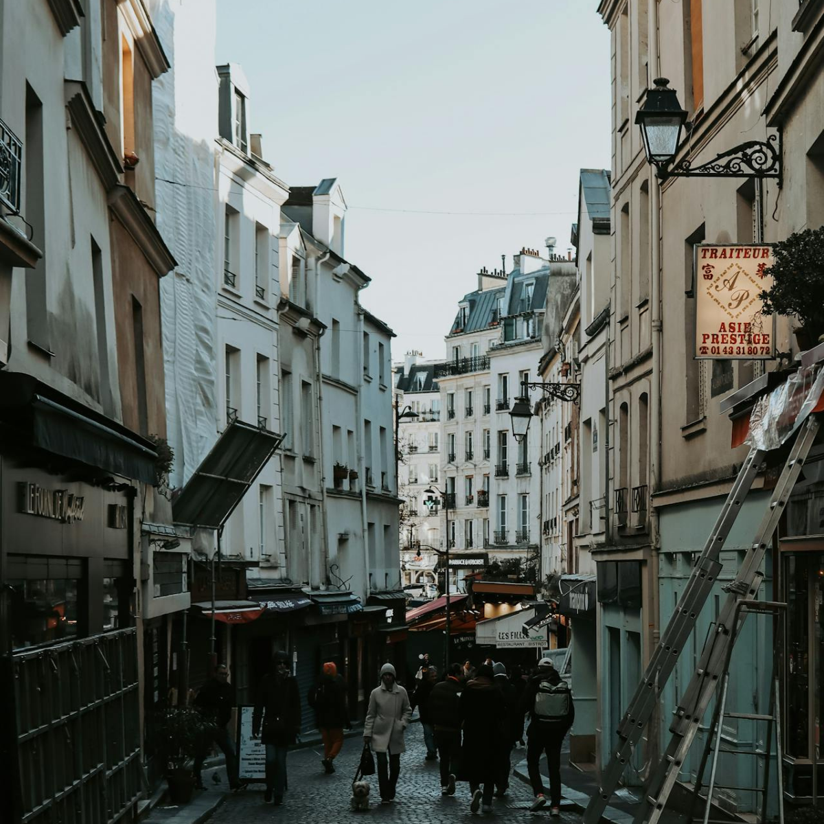 Narrow Parisian street lined with tall, cream-colored buildings. Street slopes downward; pedestrians and shops visible. 