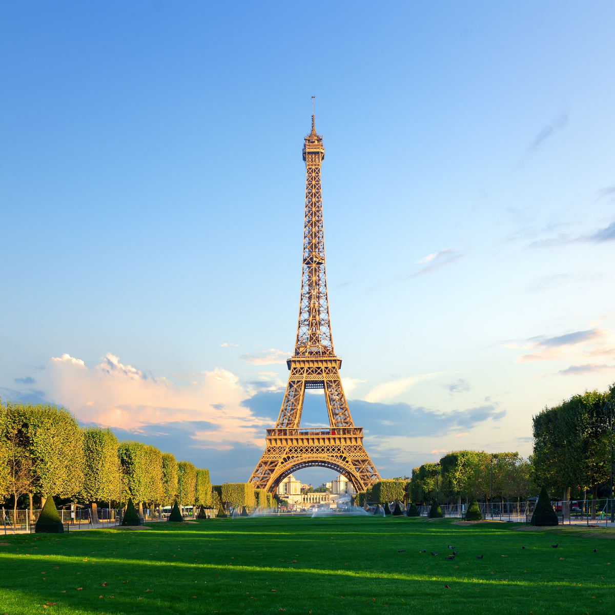 Narrow Parisian street lined with tall, cream-colored buildings. Street slopes downward; pedestrians and shops visible. 