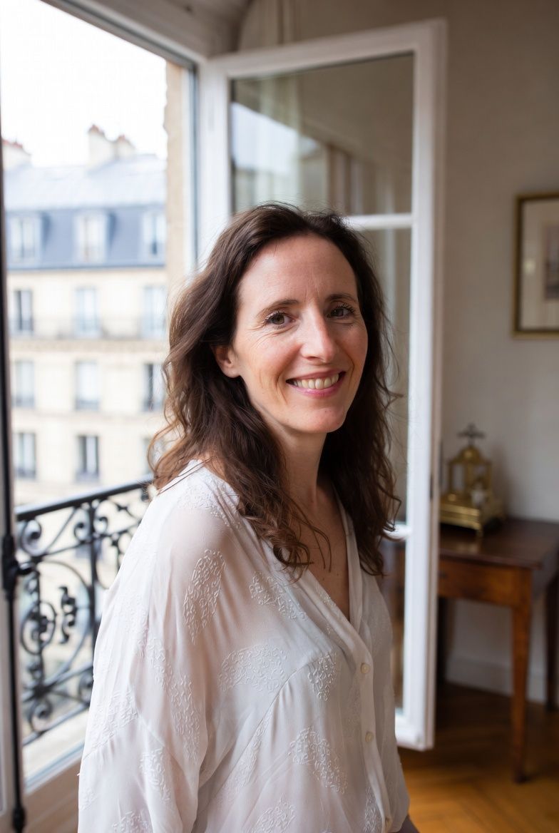 Woman with glasses smiles, black shirt, against white wall, plant in the background.
