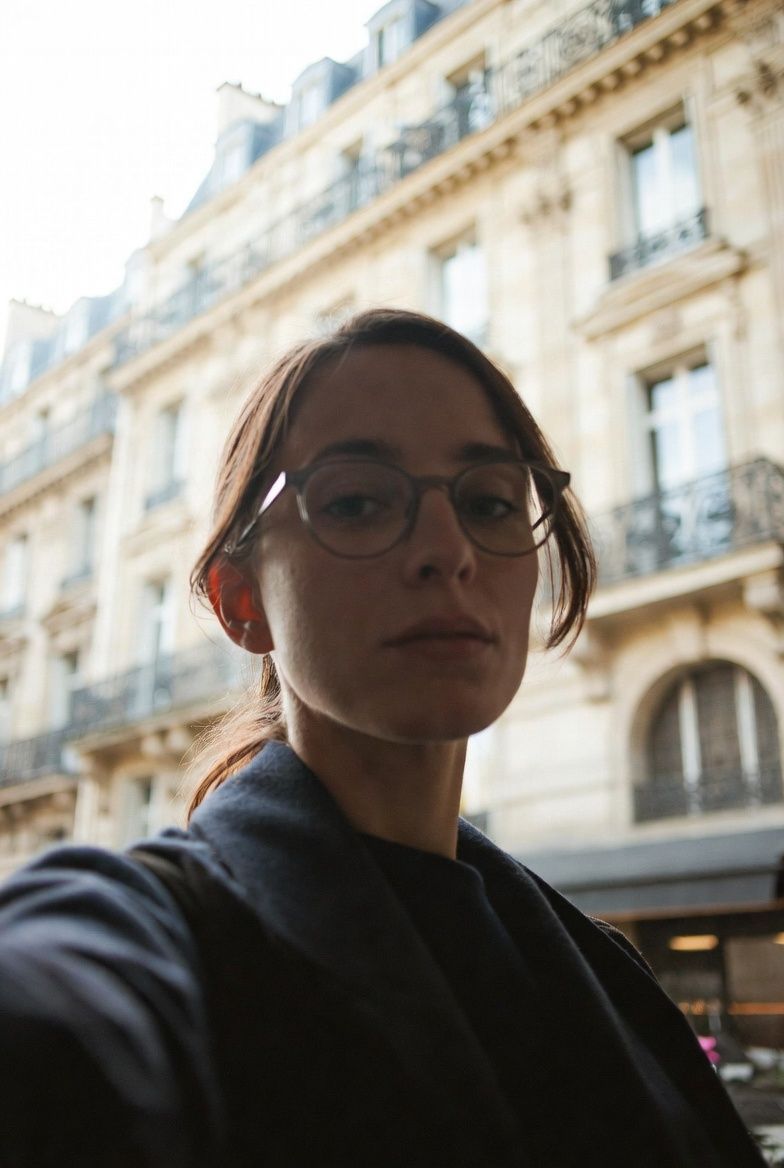 Woman laughs in front of a carousel. She wears a brown coat and sits on a bench. The setting is outdoors.
