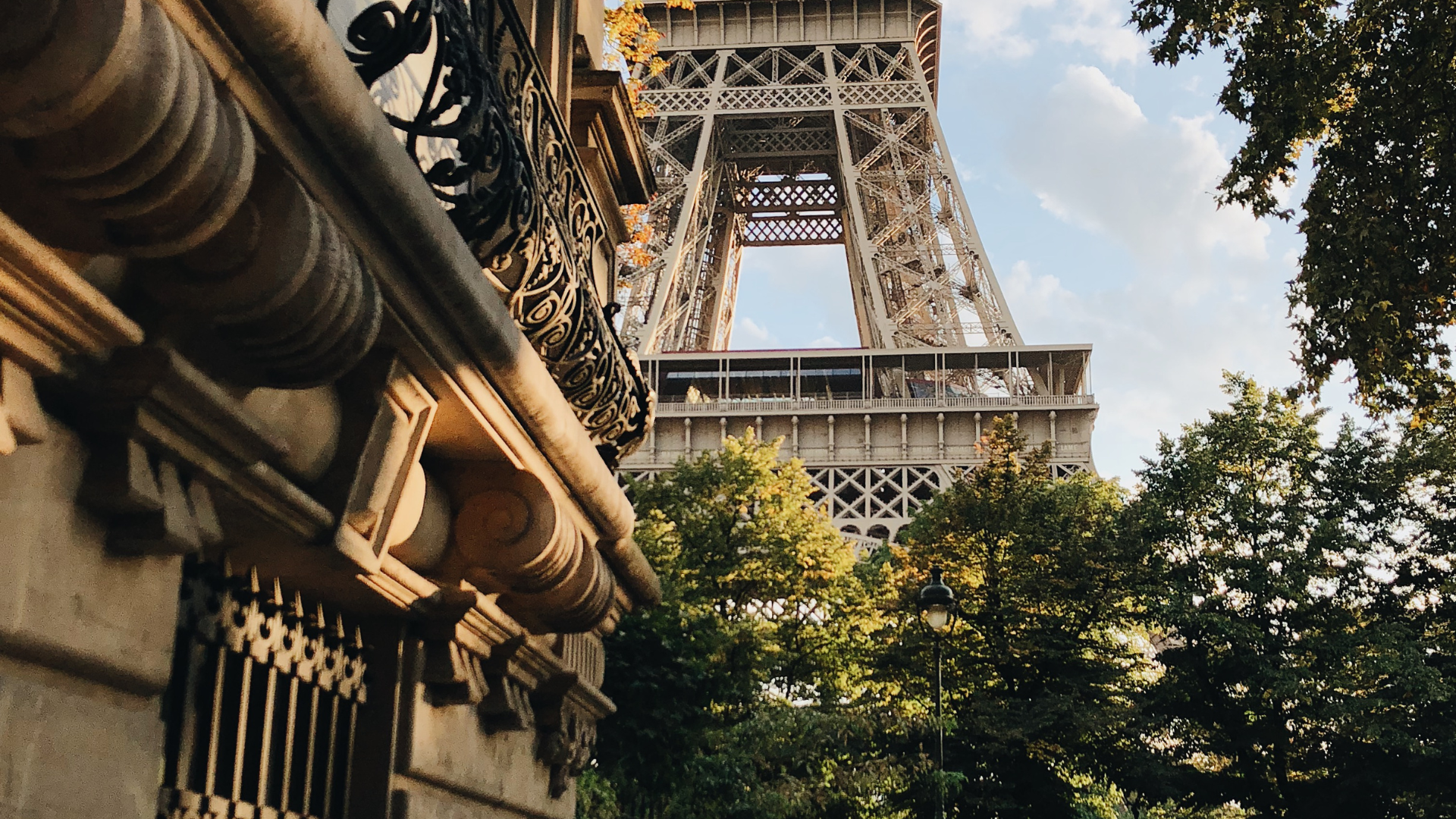 Eiffel Tower rises behind a building's stone facade and trees in Paris, France, under a blue sky.