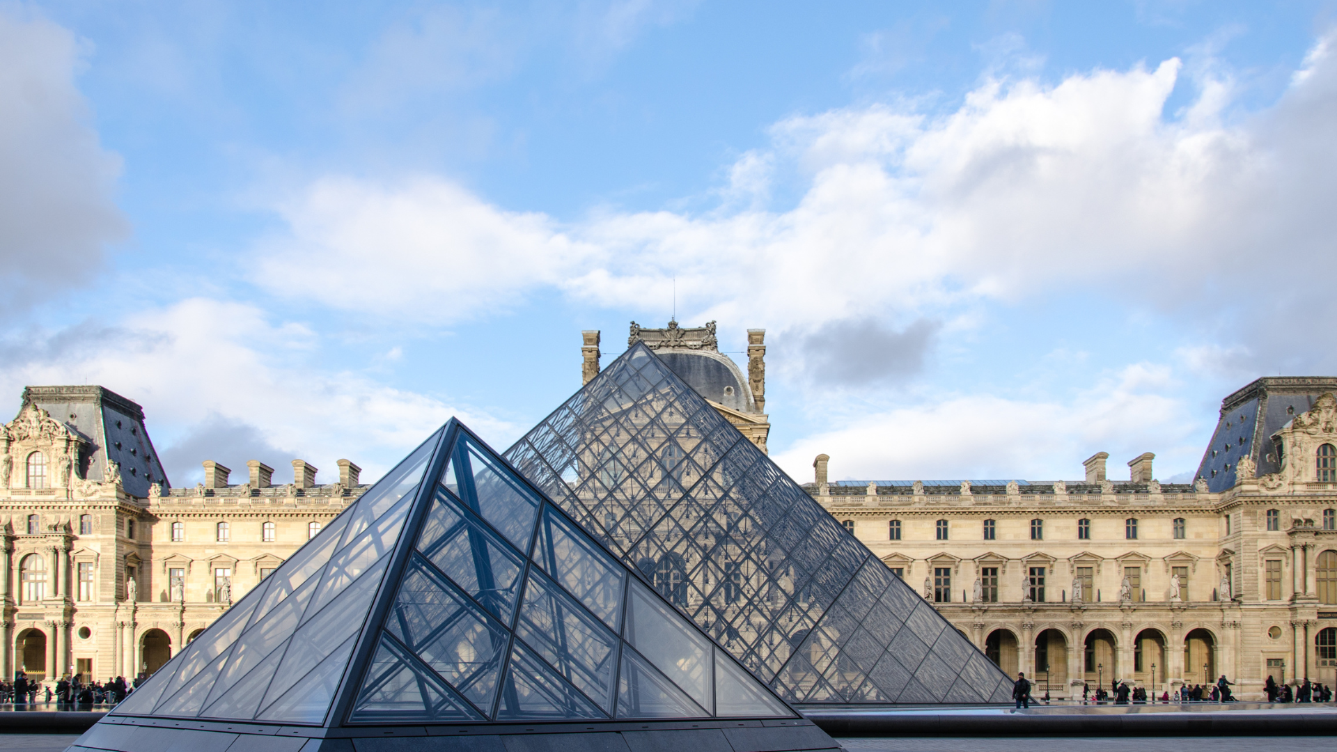 The Louvre Museum in Paris, France, with the iconic glass pyramids in the foreground. The architecture is classical, and the sky is partly cloudy.