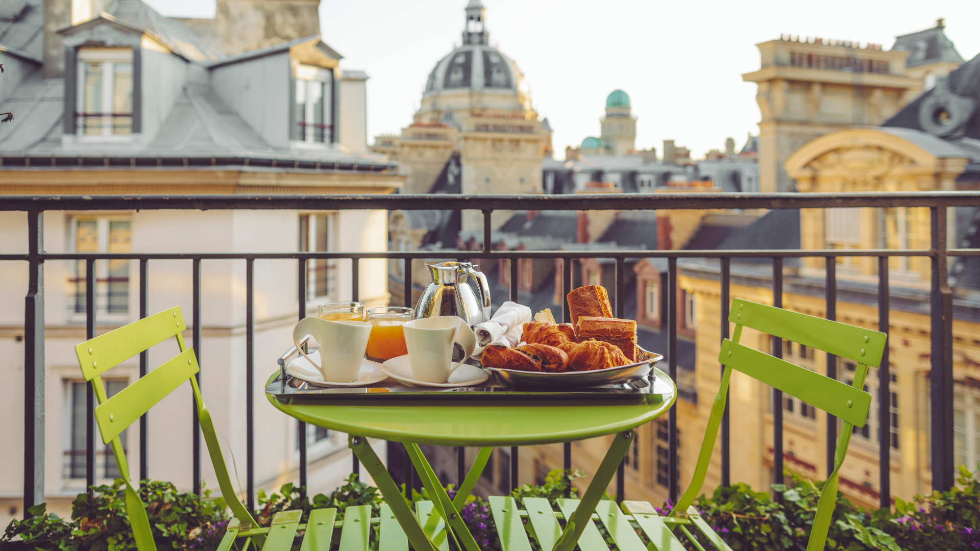 Breakfast on a Parisian balcony: green table, tray with food, city buildings in background.