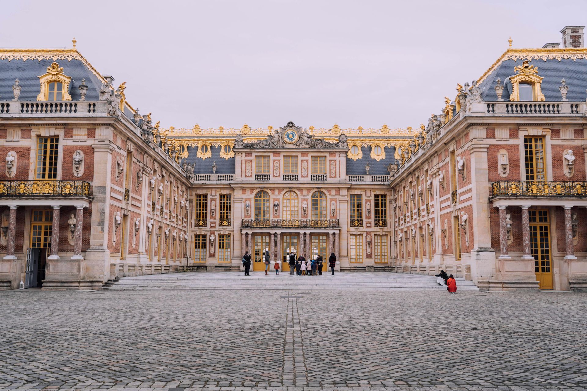 Exterior view of the Palace of Versailles, France, with a cobblestone courtyard and ornate gold and red facade.