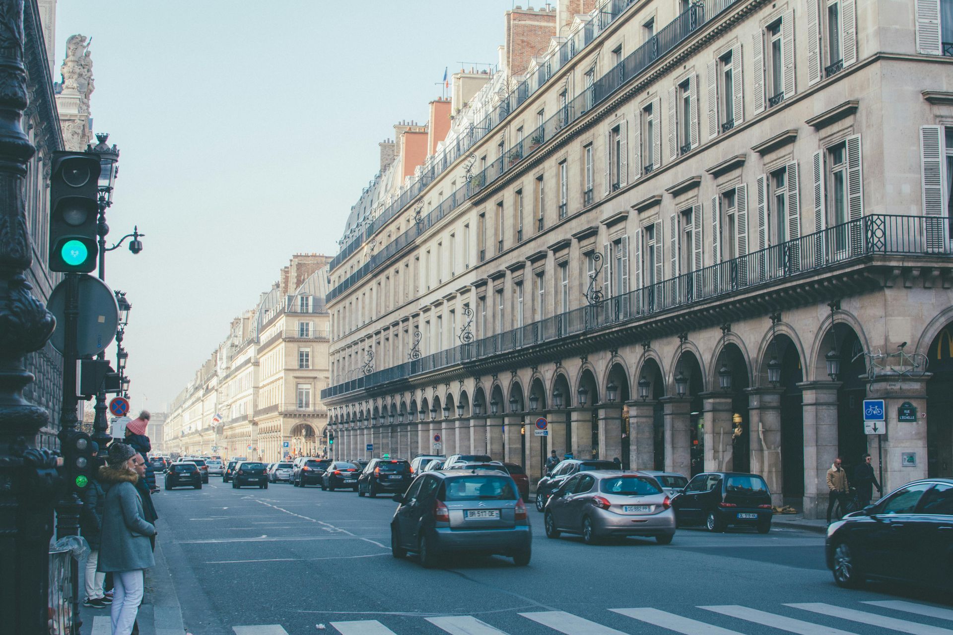 Street scene in Paris with cars, pedestrians, and ornate buildings with balconies and arched entryways.