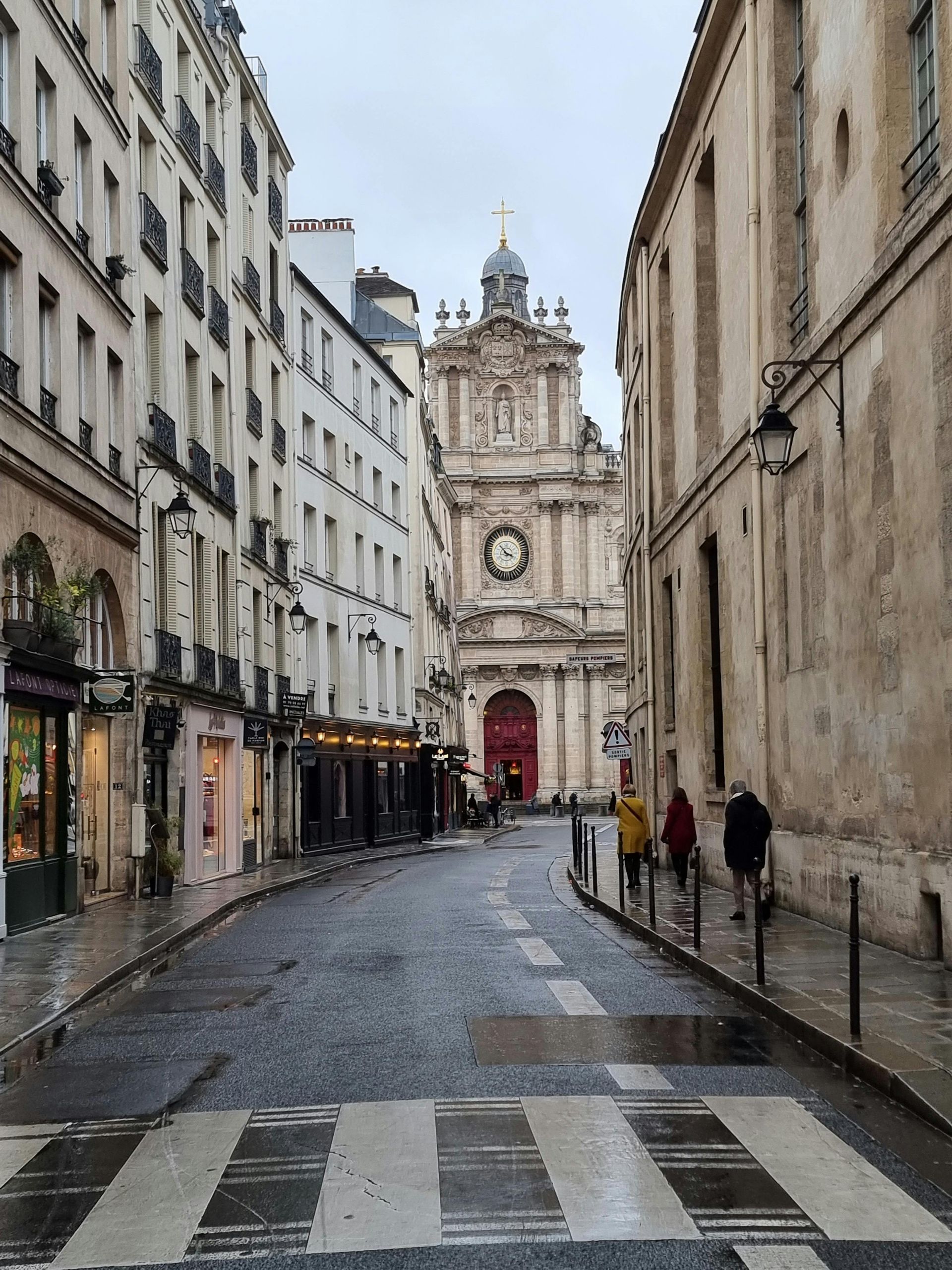 Cobblestone street in Paris leading to a grand church with a red door. Buildings line both sides.