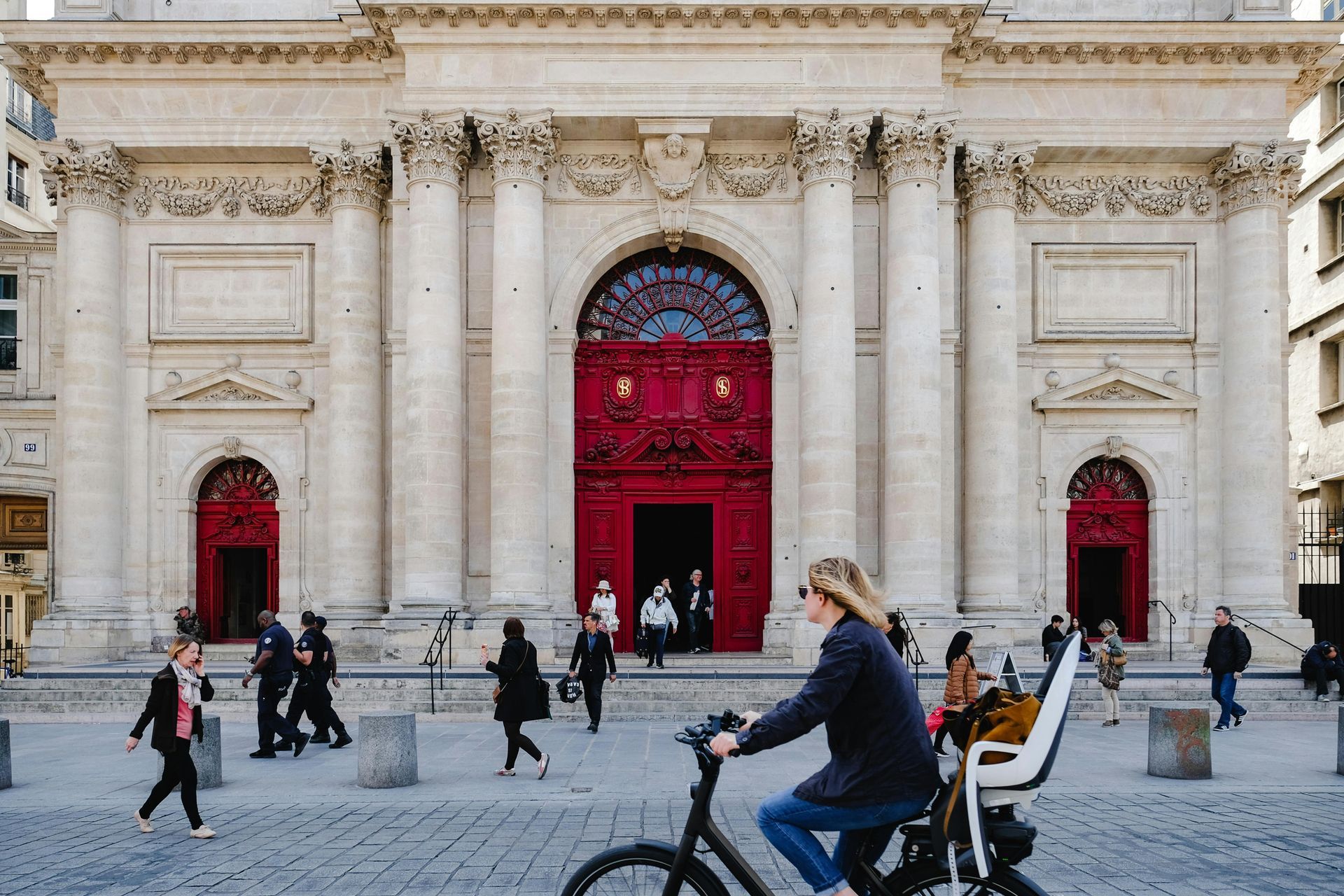 People walking and biking in front of a classical building with red doors and columns in a cobblestone square.