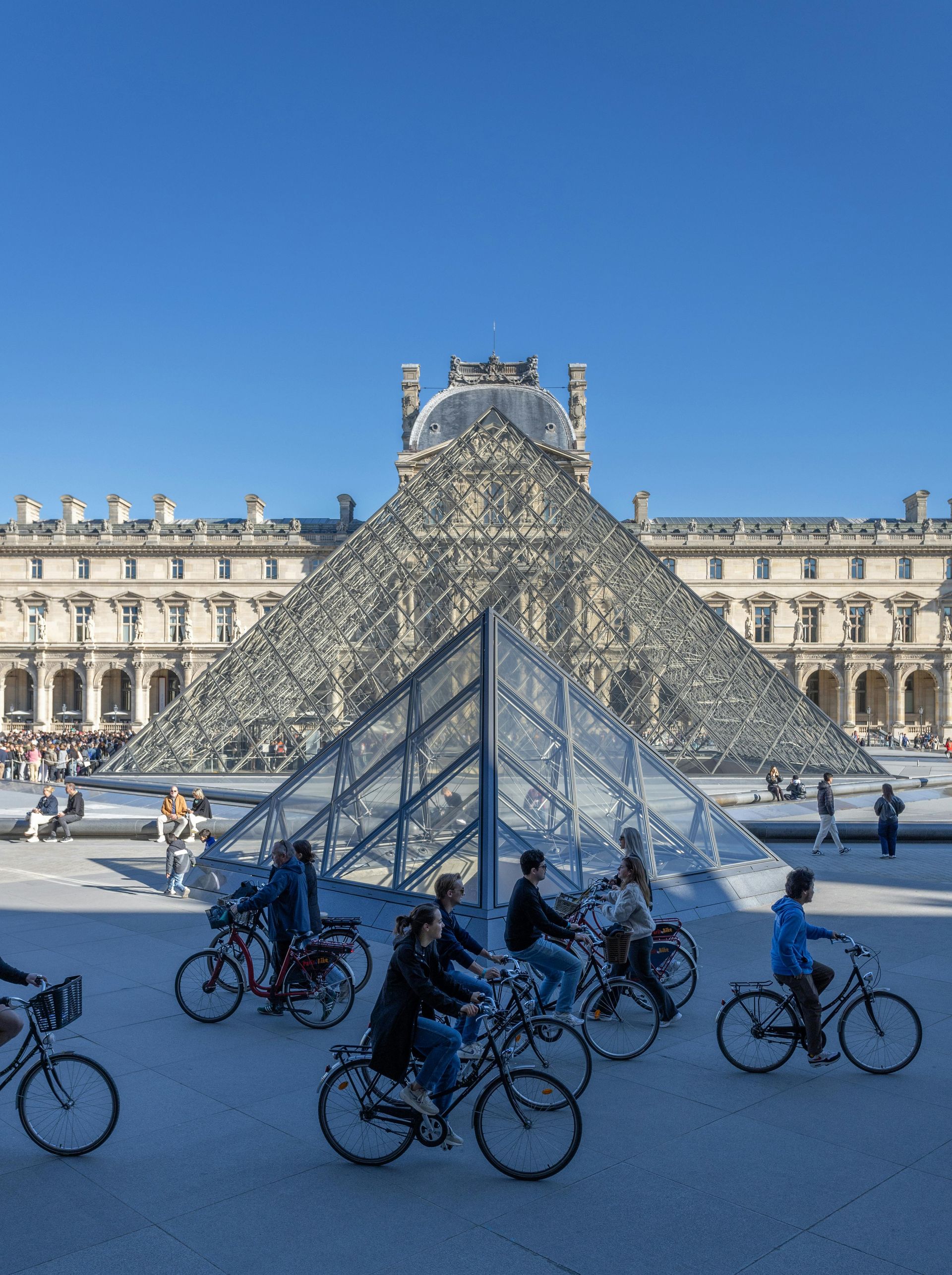 Cyclists in front of the Louvre Pyramid in Paris, France, on a sunny day.