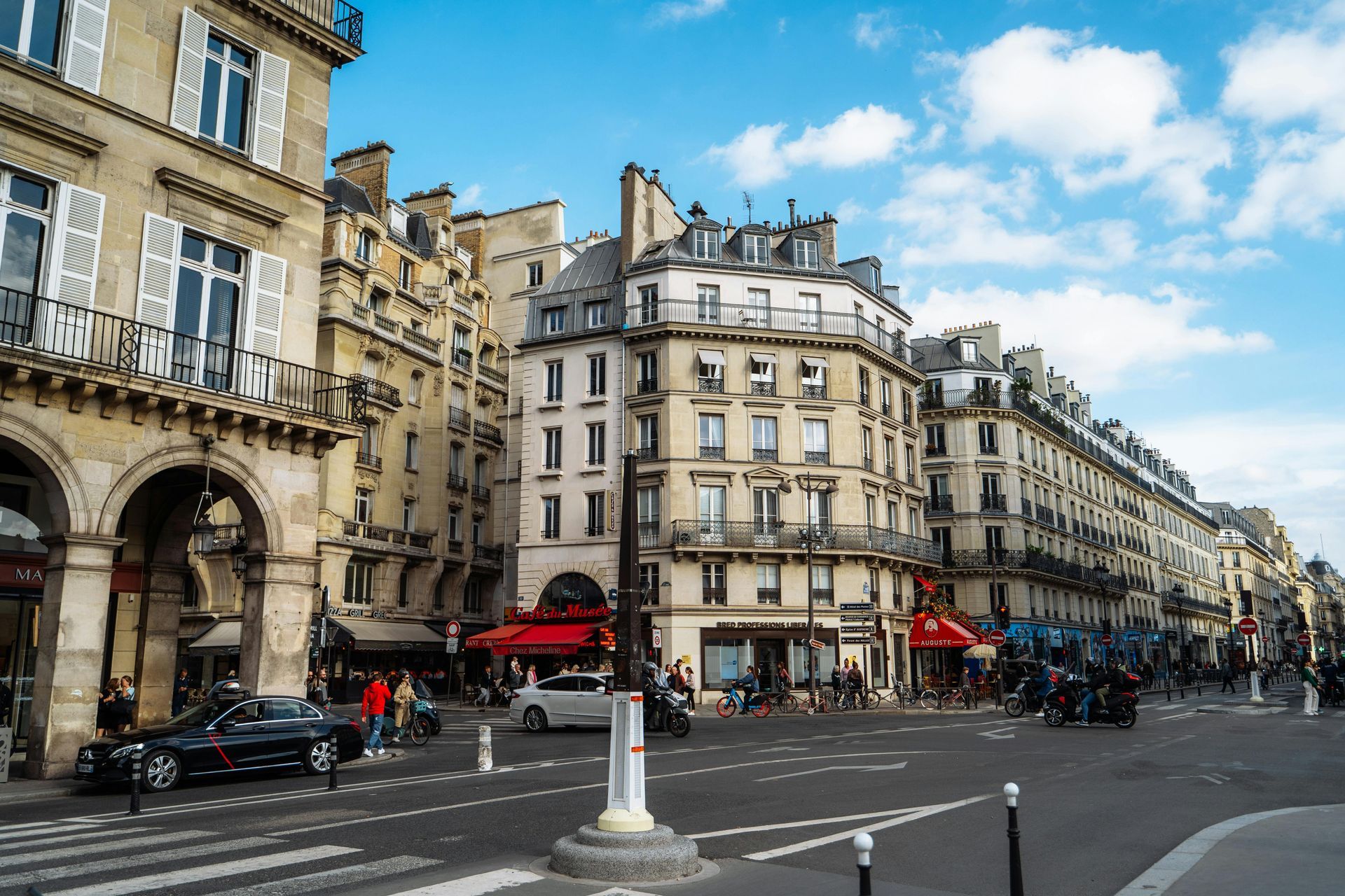 Parisian street scene featuring buildings with balconies, shops, traffic, and pedestrians under a blue sky.