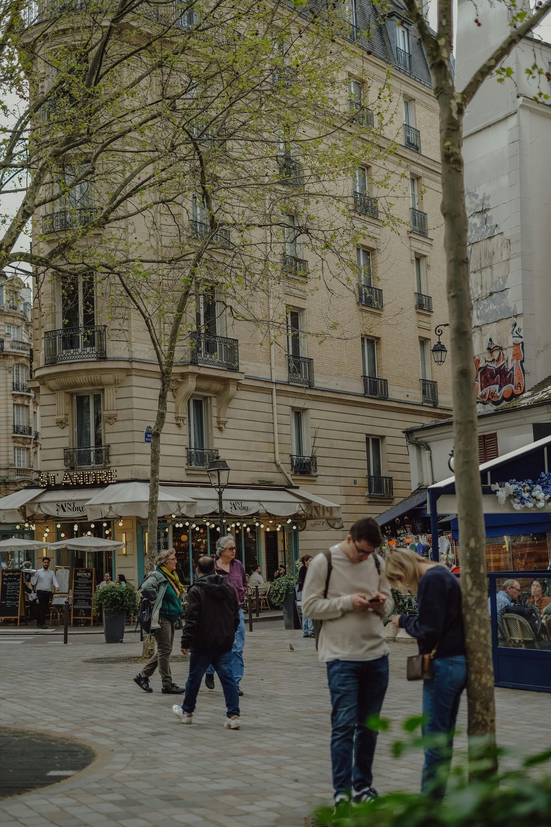 People walking near a beige building with balconies and a cafe, in an outdoor Parisian setting.