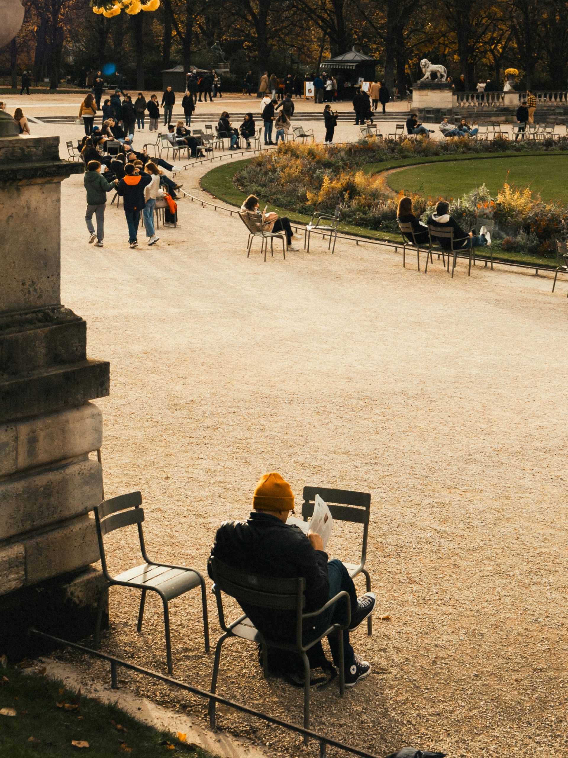 Man in orange hat reads newspaper in a park. People stroll on a gravel path; trees in the background.