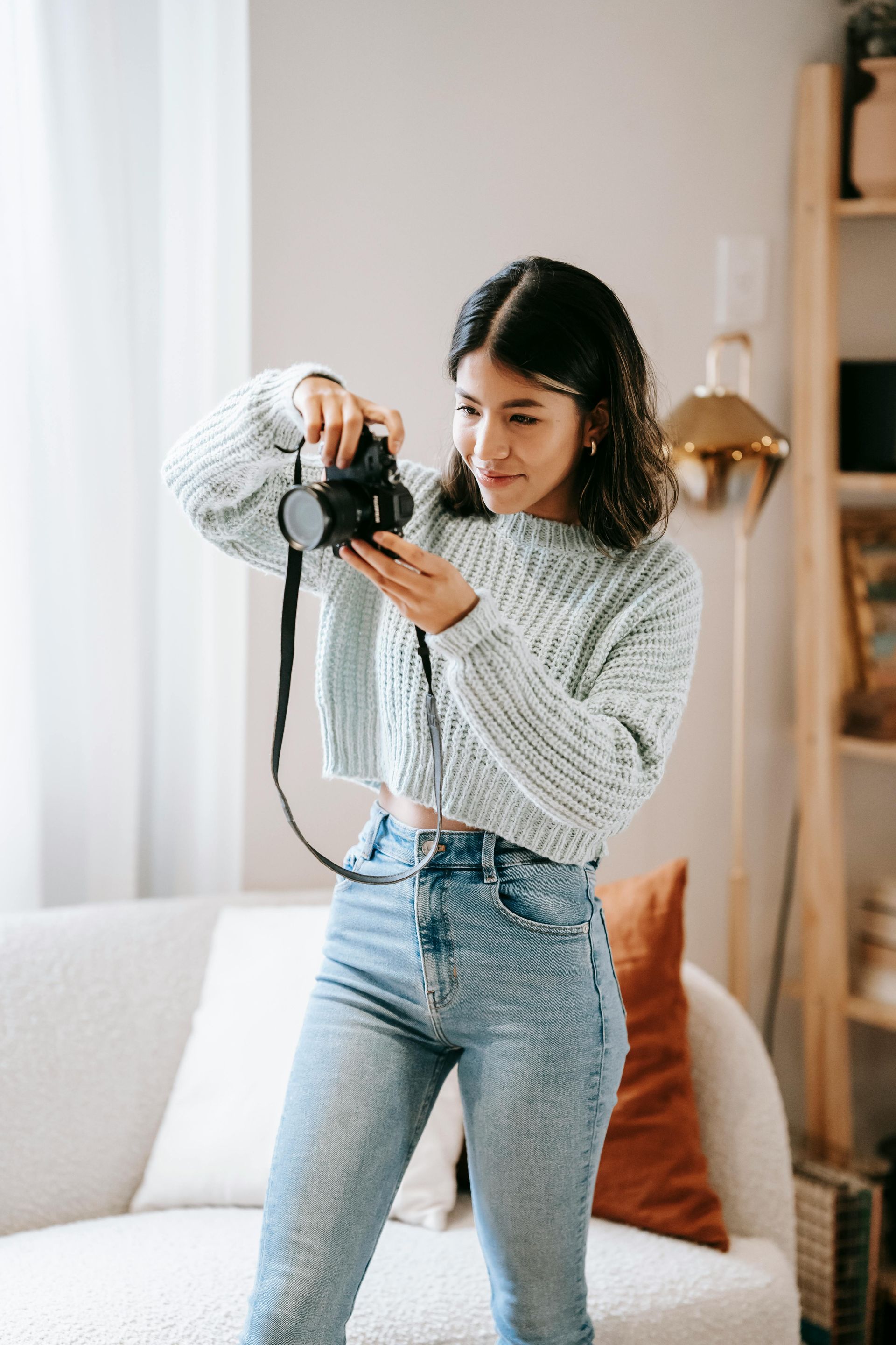 Woman in light blue sweater and jeans taking a photo with a black camera in a living room.