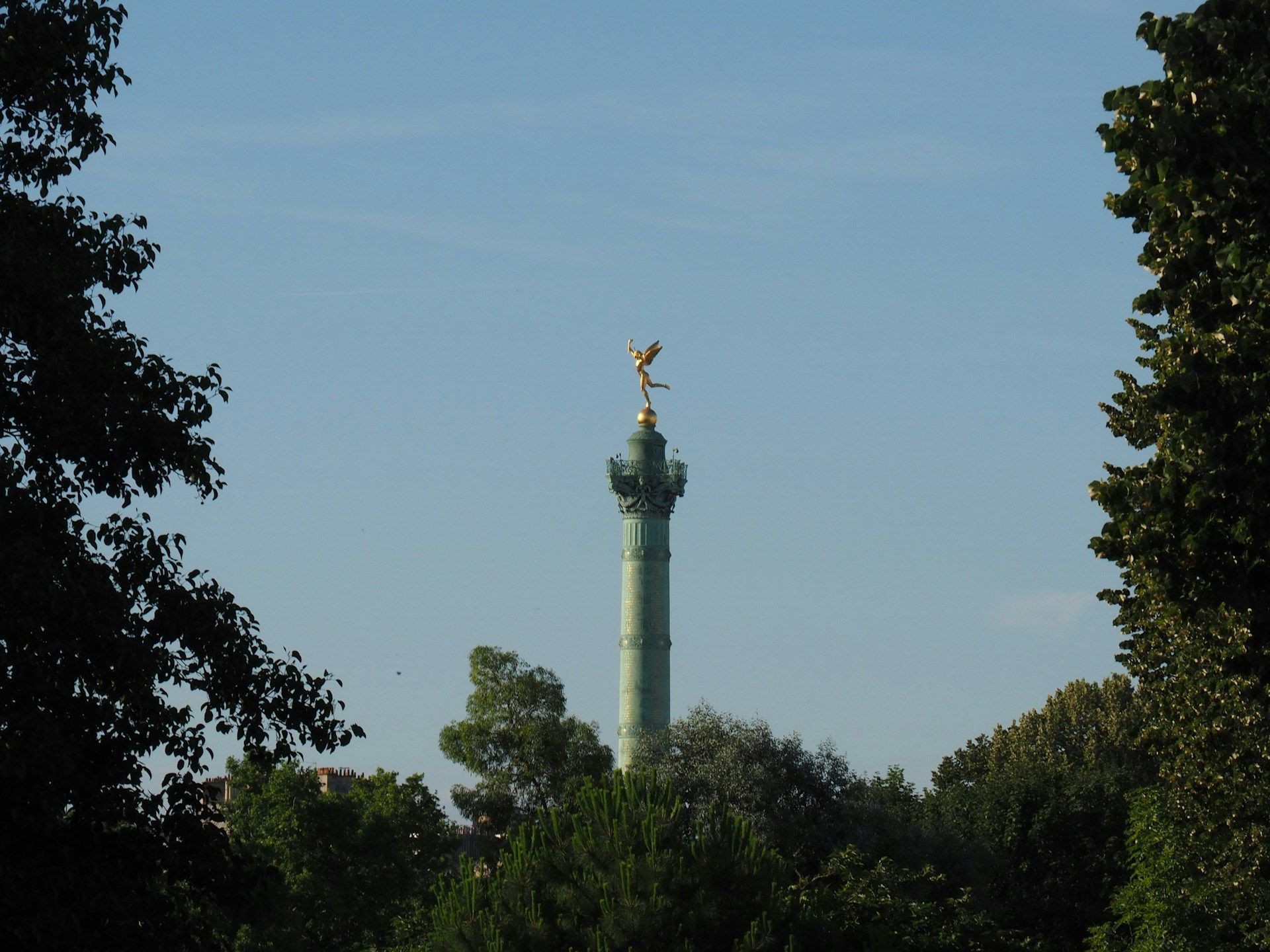 Column topped with golden statue against blue sky, framed by tree branches.