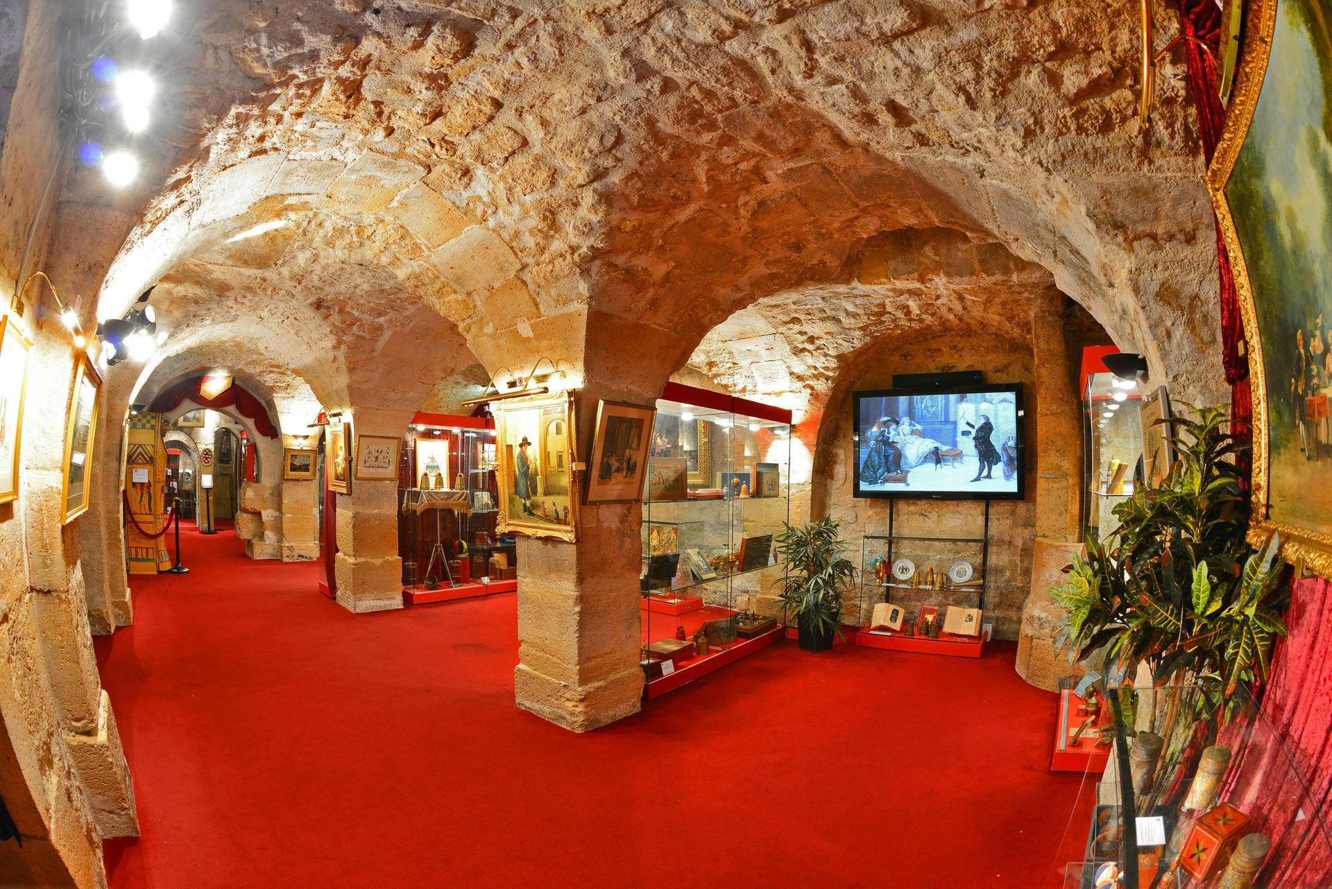 Museum interior with red carpet, stone columns and ceiling, displaying art and a video screen.
