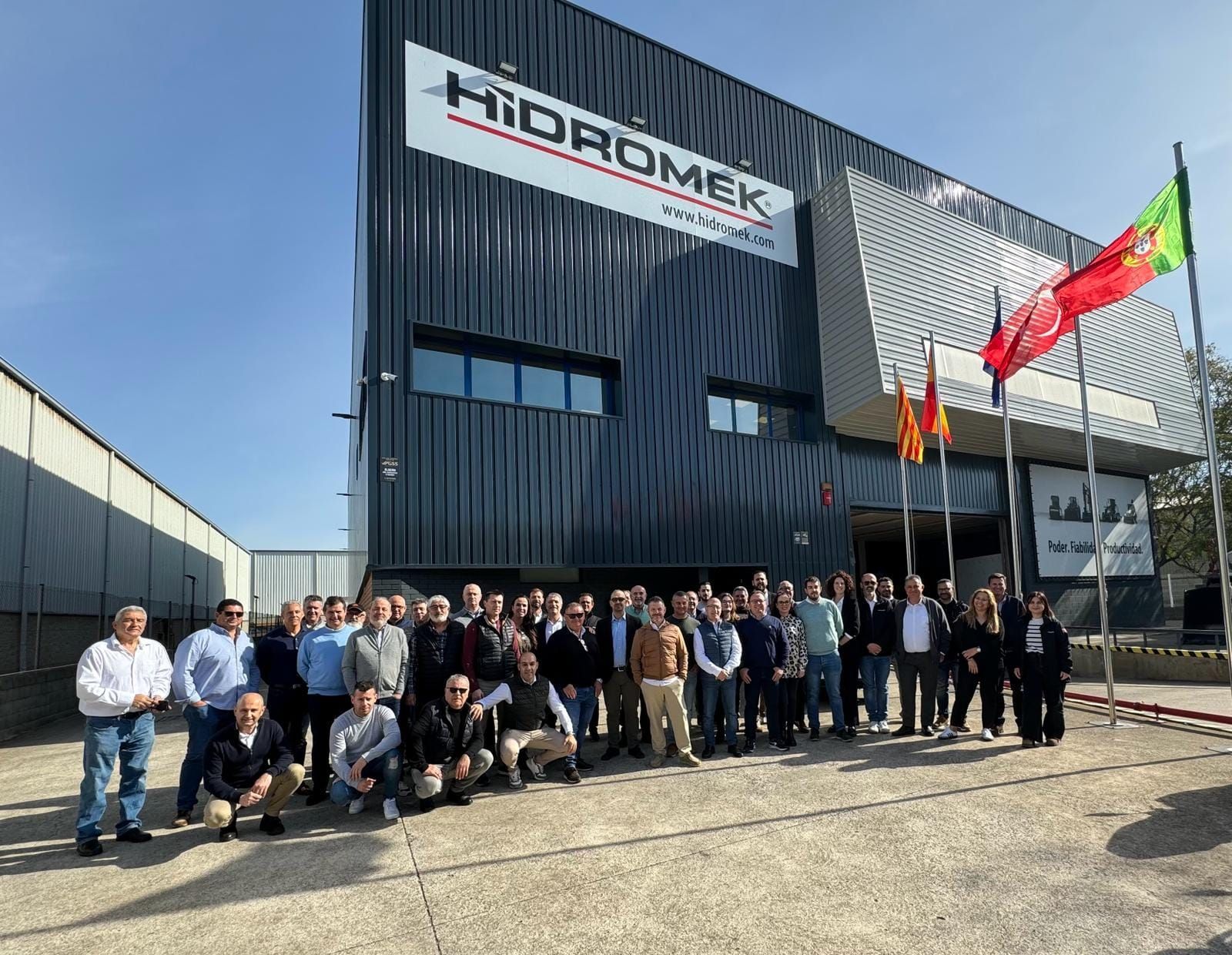 Grupo de personas posando frente al edificio de la fábrica HIDROMEK en un día soleado, con banderas en primer plano.