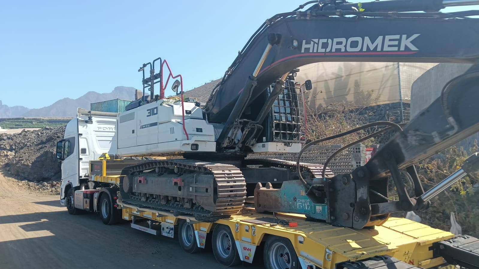 Una excavadora pesada cargada en un remolque de plataforma baja amarillo en una cantera rocosa bajo un cielo azul despejado.