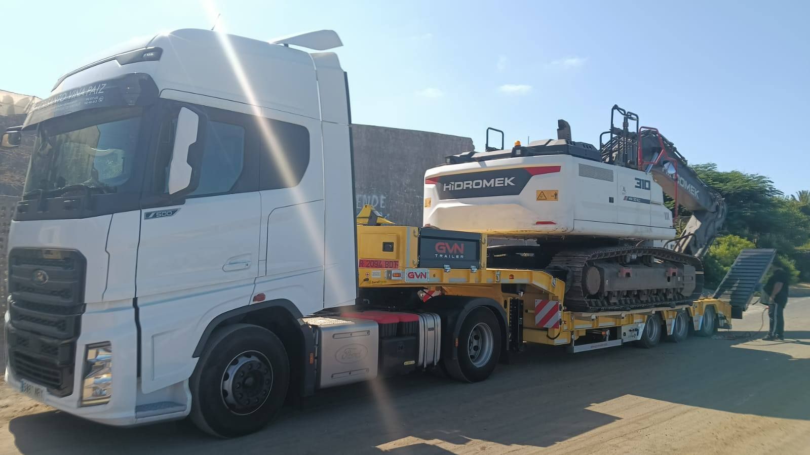 Un camión semirremolque blanco transporta una gran excavadora en un remolque de plataforma baja bajo la luz del sol.