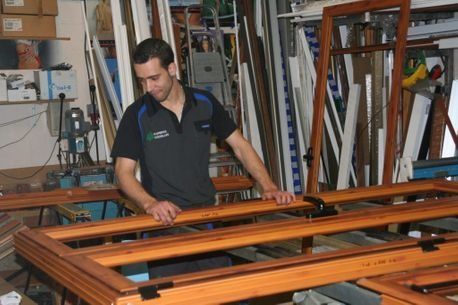 Un trabajador construye un marco de puerta de madera en un taller de carpintería lleno de tablones y herramientas.
