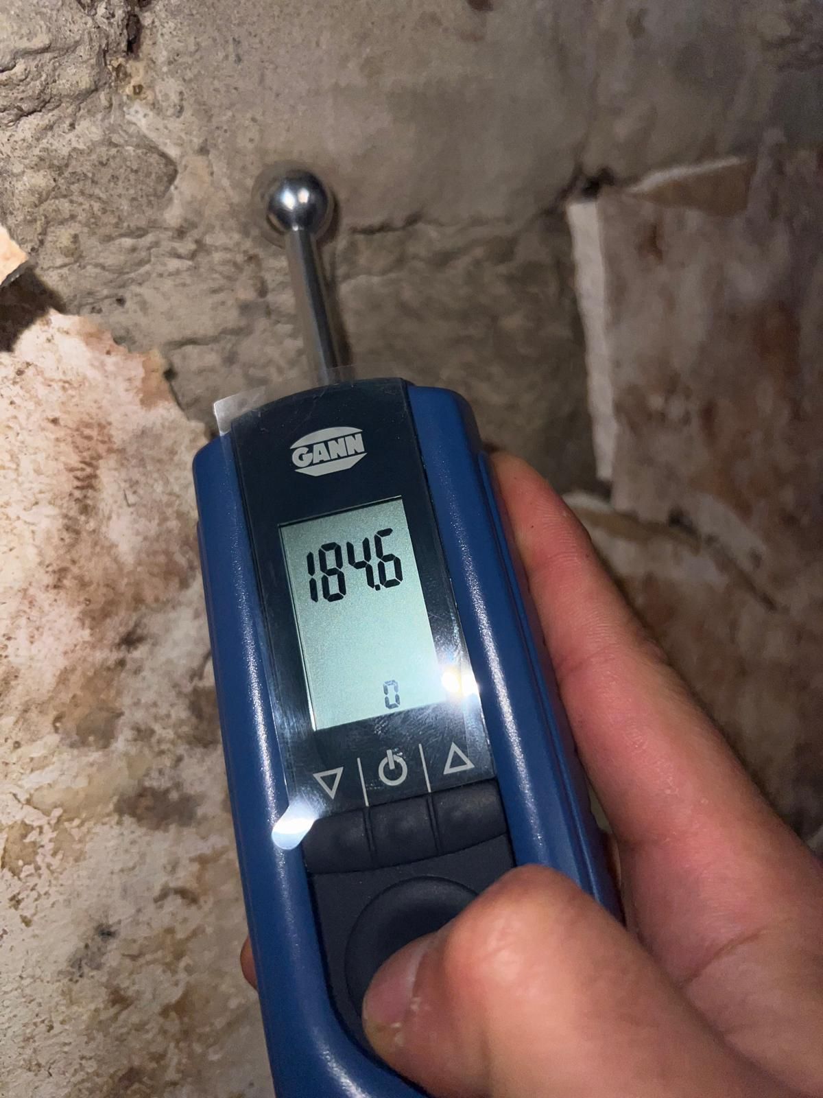 Eine Hand hält ein blaues Feuchtigkeitsmessgerät an eine Steinmauer; das Messgerät zeigt 184,6 an.
