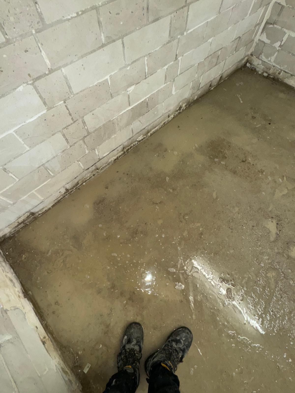Stehendes Wasser auf einem Betonboden vor Wänden aus Schlackenblöcken. Füße einer Person in schwarzen Arbeitsstiefeln.