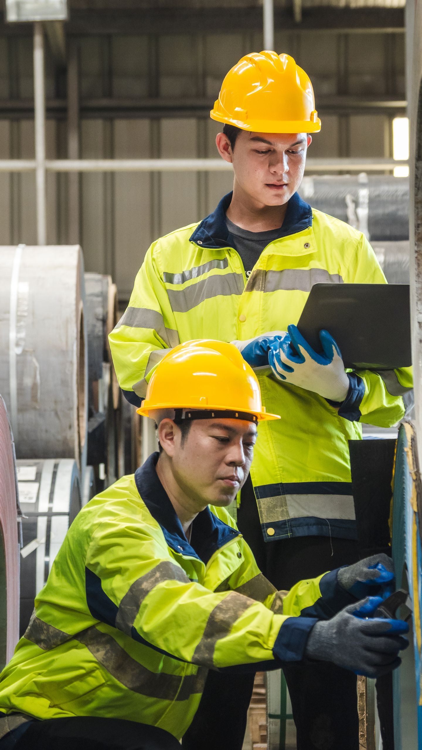 Deux ouvriers, vêtus de gilets de sécurité jaunes et de casques de chantier, inspectent des machines. L'un d'eux tient un ordinateur portable.