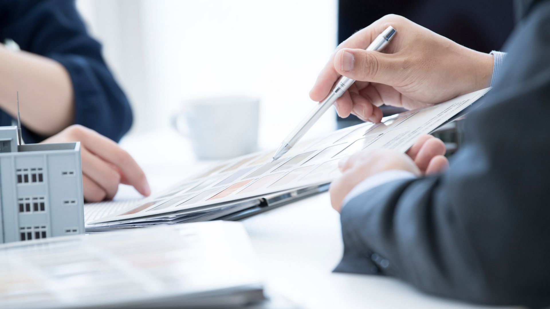 Deux personnes examinent un document, l'une d'elles le désignant avec un stylo. Une tasse blanche et une petite maquette de bâtiment sont posées sur la table.