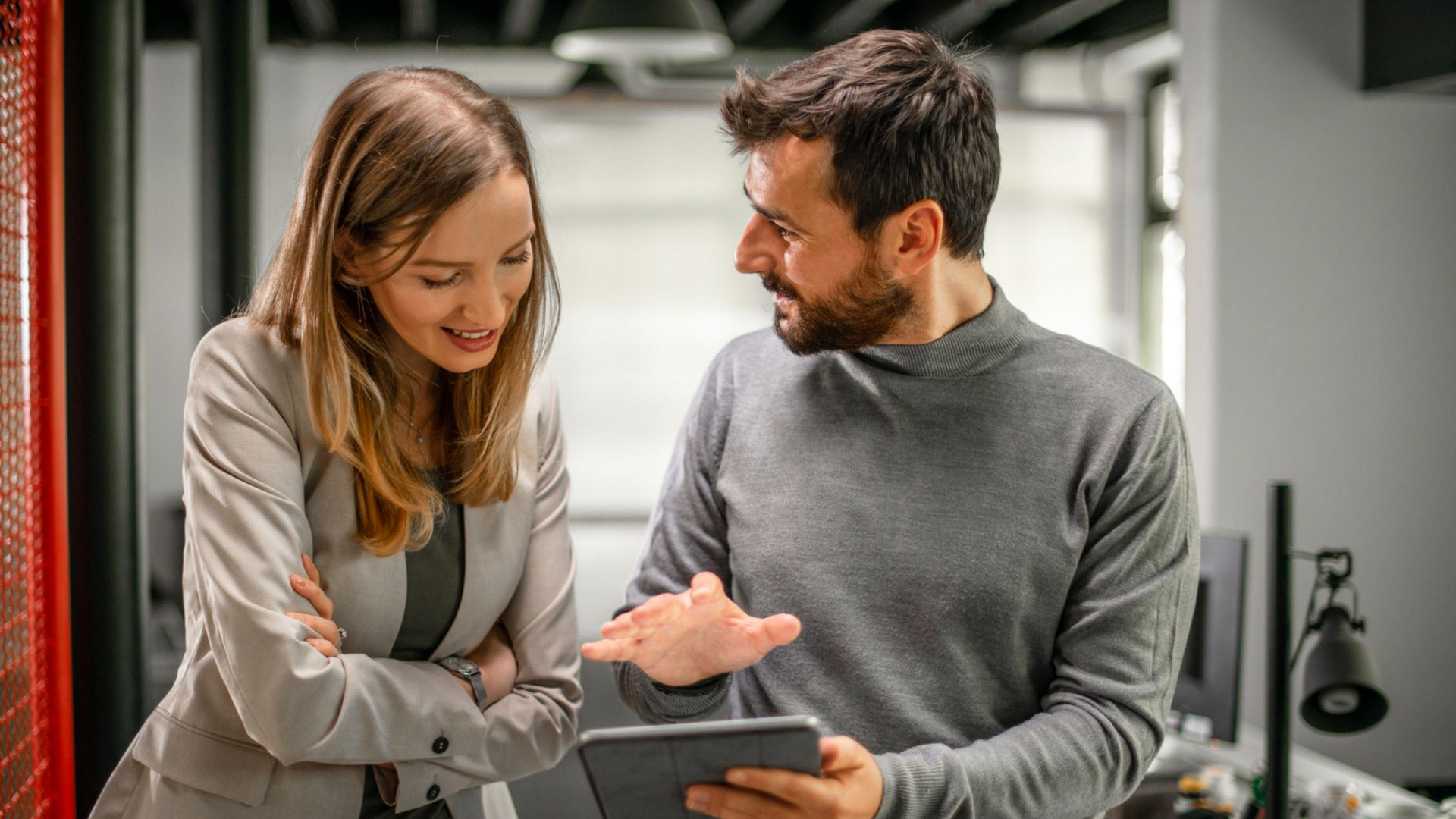Une femme, les bras croisés, rit tandis qu'un homme désigne une tablette ; contexte de bureau.