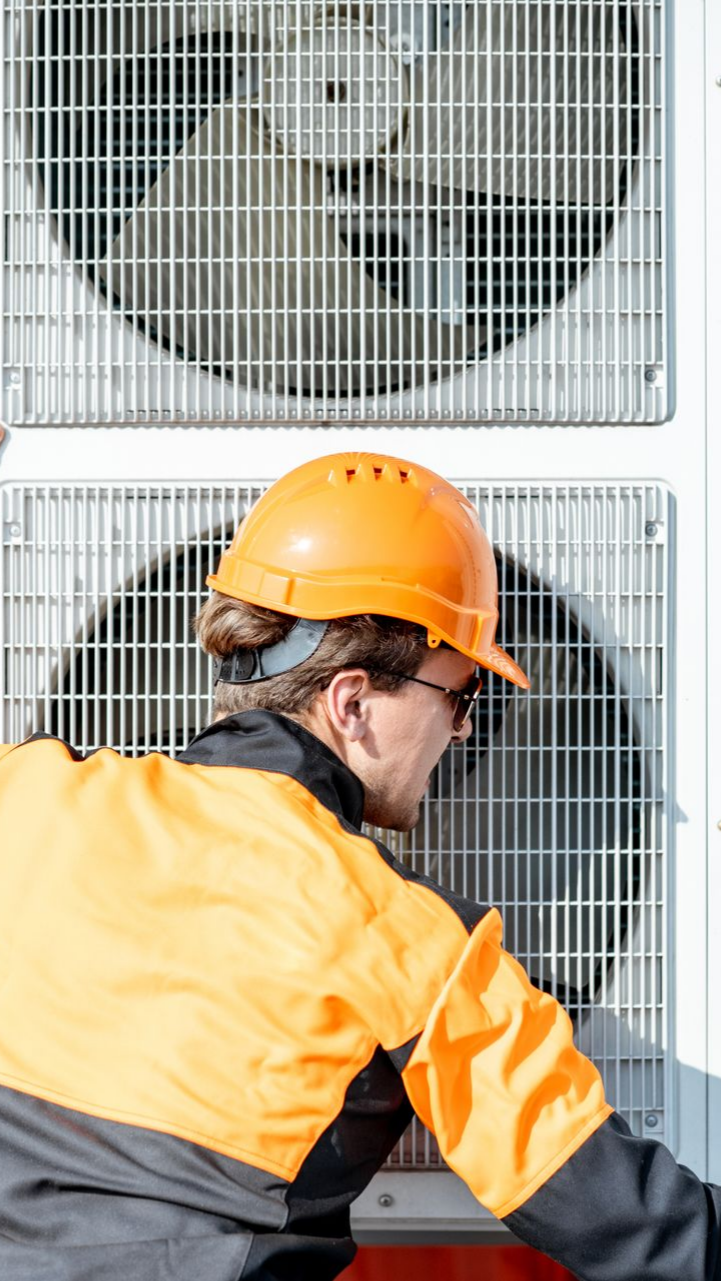 Une personne portant un casque de chantier orange et une veste de sécurité inspecte un climatiseur.