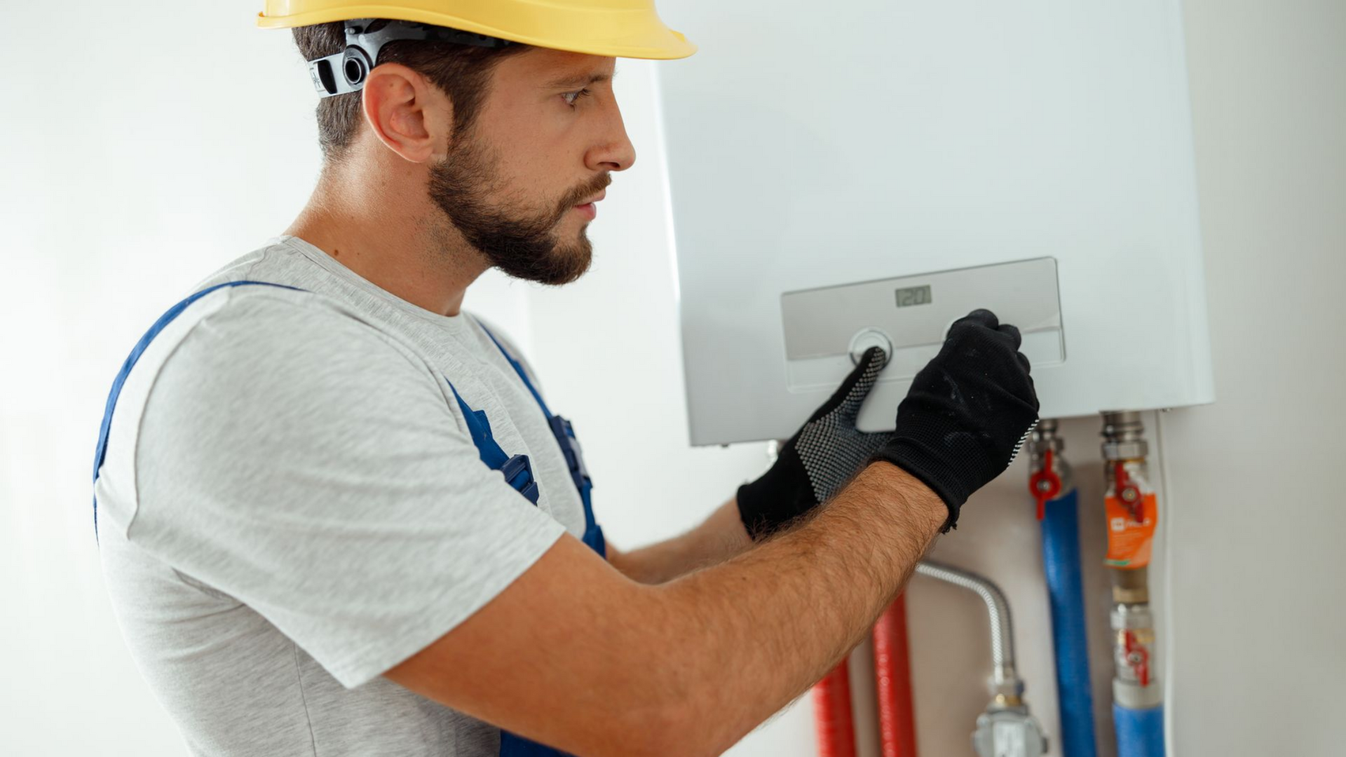 Un homme en tenue de travail ajuste un radiateur mural blanc. Il porte un casque et des gants ; on aperçoit les tuyaux.