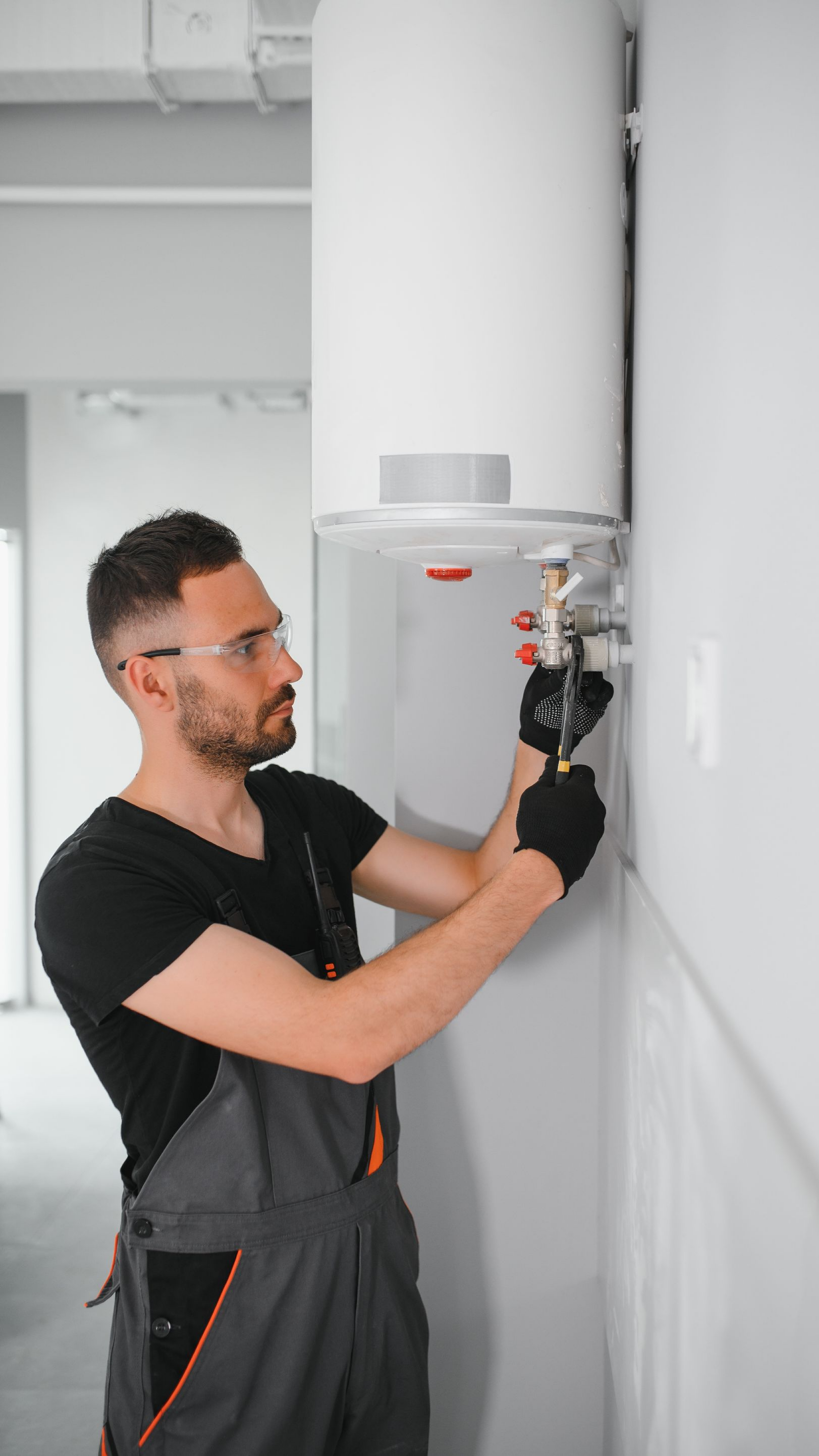 Un plombier en salopette et lunettes de sécurité travaille sur un chauffe-eau, à l'intérieur.