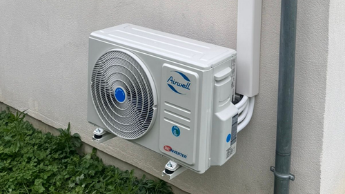 Unité de climatisation blanche fixée sur un mur extérieur clair, avec une petite parcelle d'herbe verte en dessous.