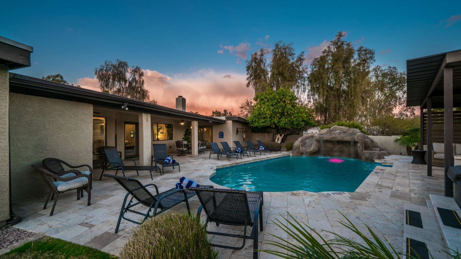 Backyard with swimming pool, lounge chairs, and a rock waterfall at sunset.