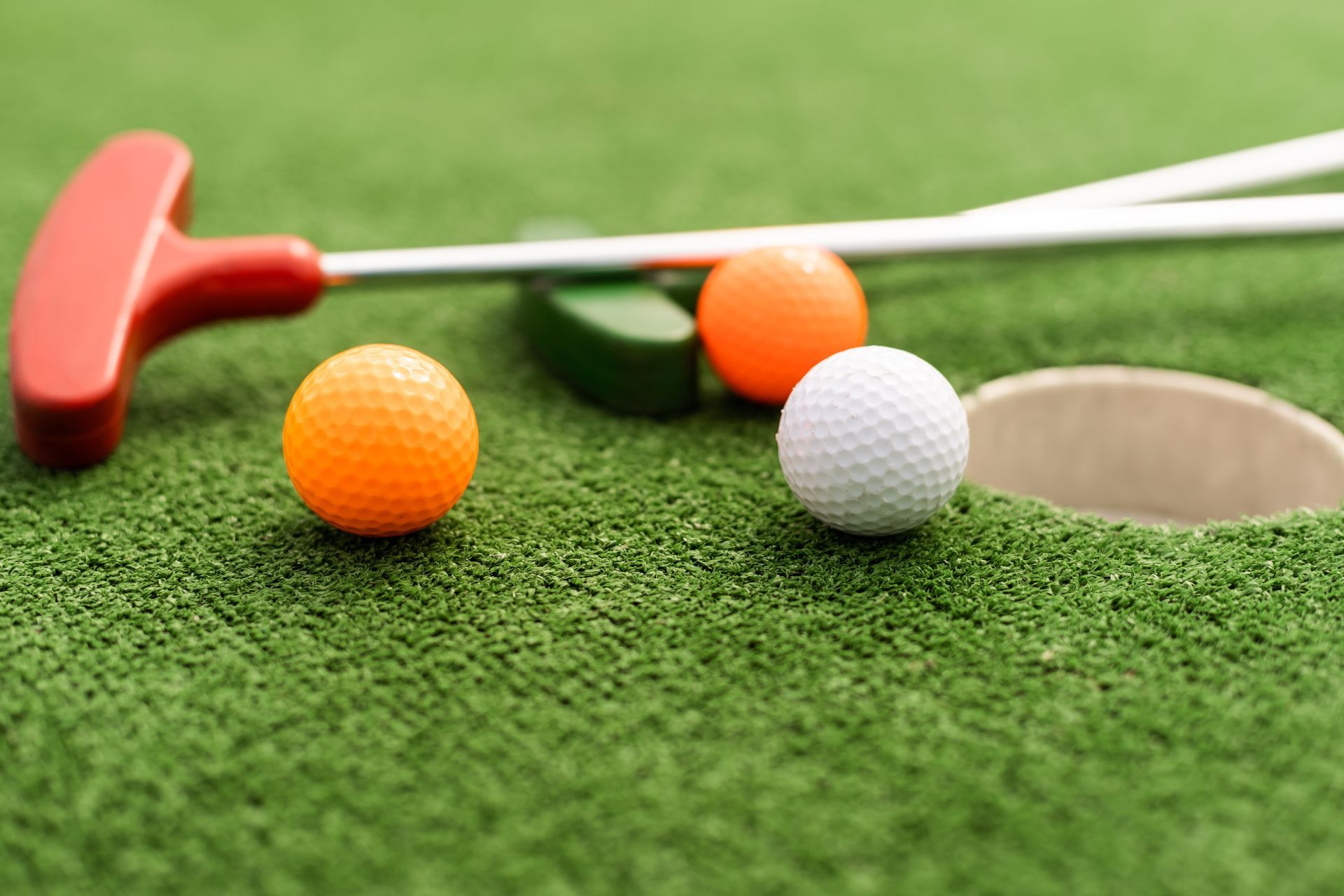 Miniature golf setup with orange and white golf balls near a hole and putter on green turf.