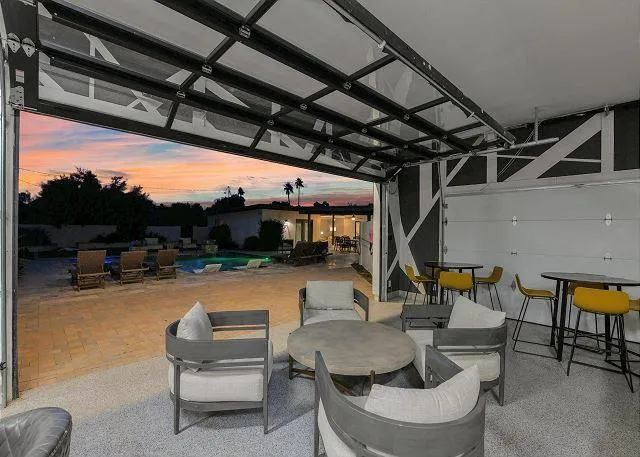 Patio with open glass garage door, seating, pool in the background at dusk.