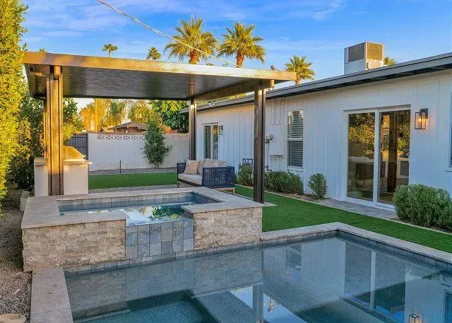 Backyard with pool, hot tub, seating area under a pergola, and white house. Palm trees in the background.