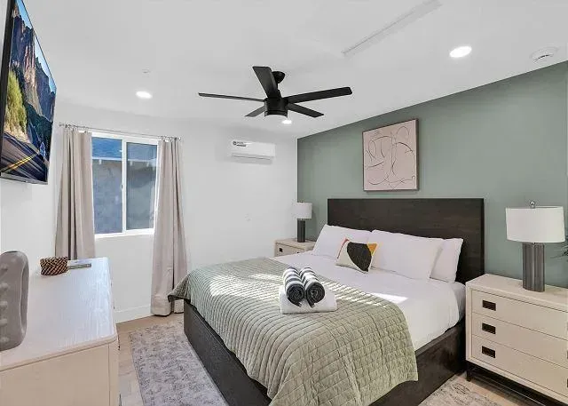 Bedroom with queen bed, mint green accent wall, white dresser, large TV, ceiling fan, and window.
