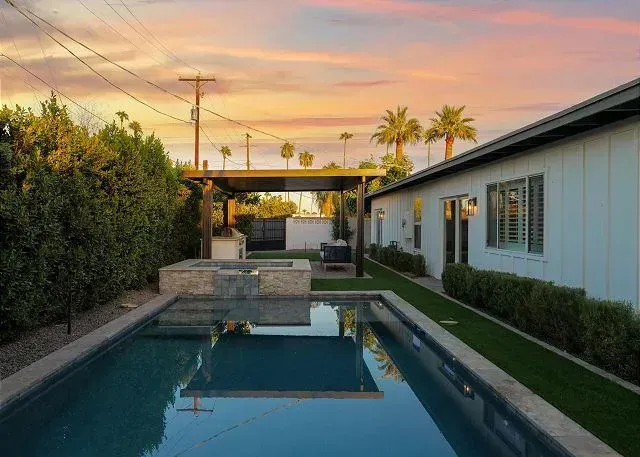 Backyard with pool, outdoor kitchen under pergola, white house, sunset sky, palm trees.