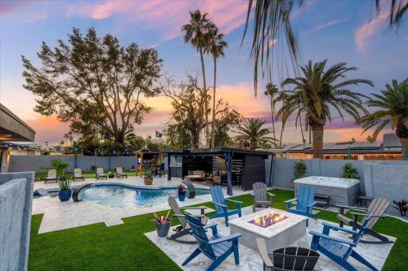 Backyard with a pool, hot tub, fire pit, and seating under a colorful sunset. Palm trees in the background.